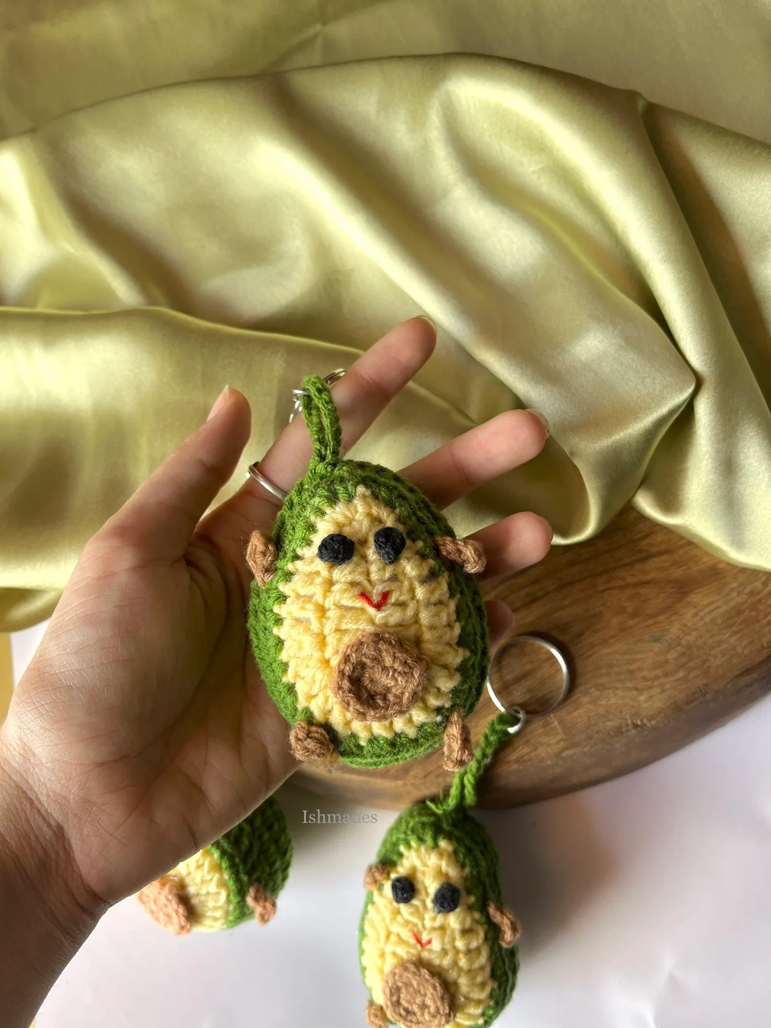 Hand holding a crochet avocado keychain with cute face detail, wooden board in background.