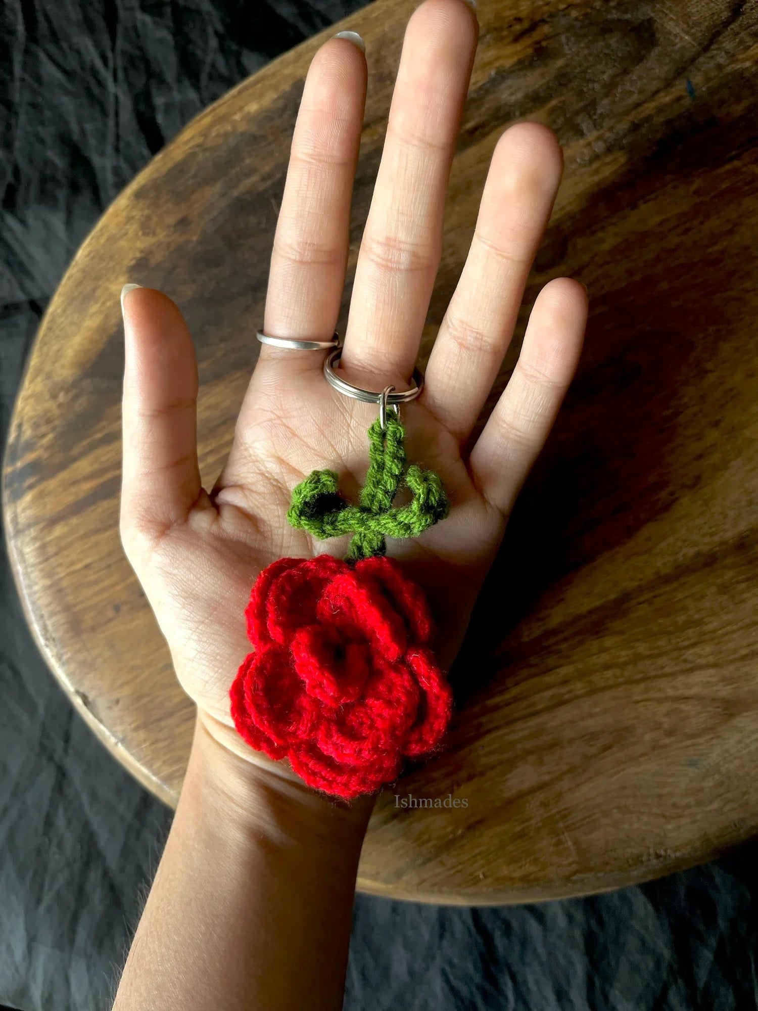 Hand holding a red crochet rose keychain with green leaves on a wooden table