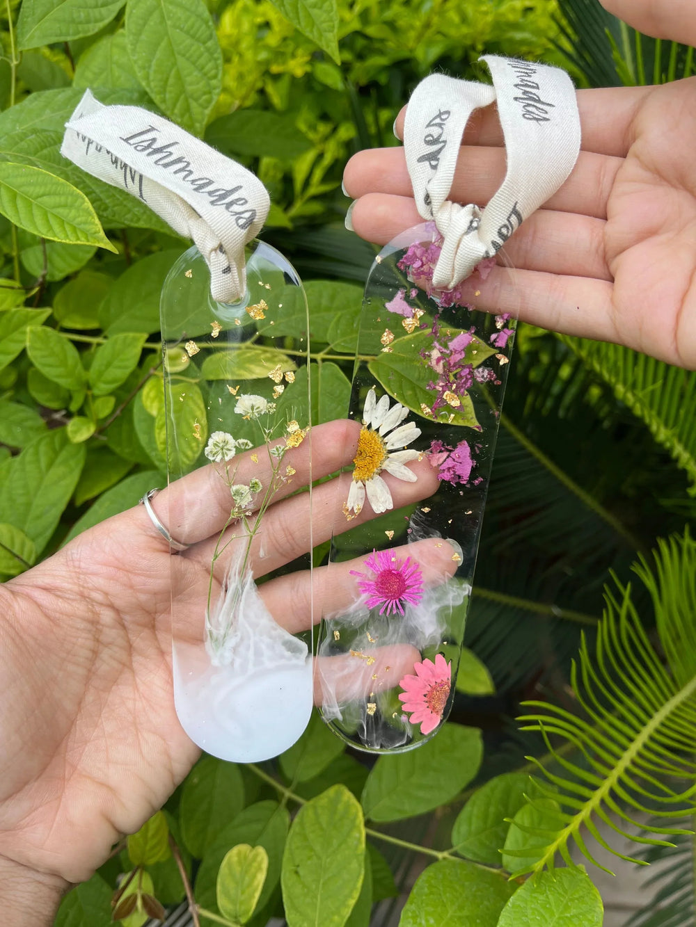 Hand holding two floral resin bookmarks with dried flowers and ribbon, outdoors with green leaves