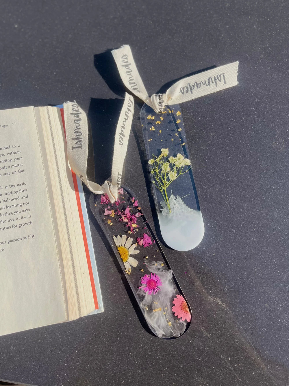 Resin bookmarks with pressed flowers and gold flakes on a dark surface beside an open book