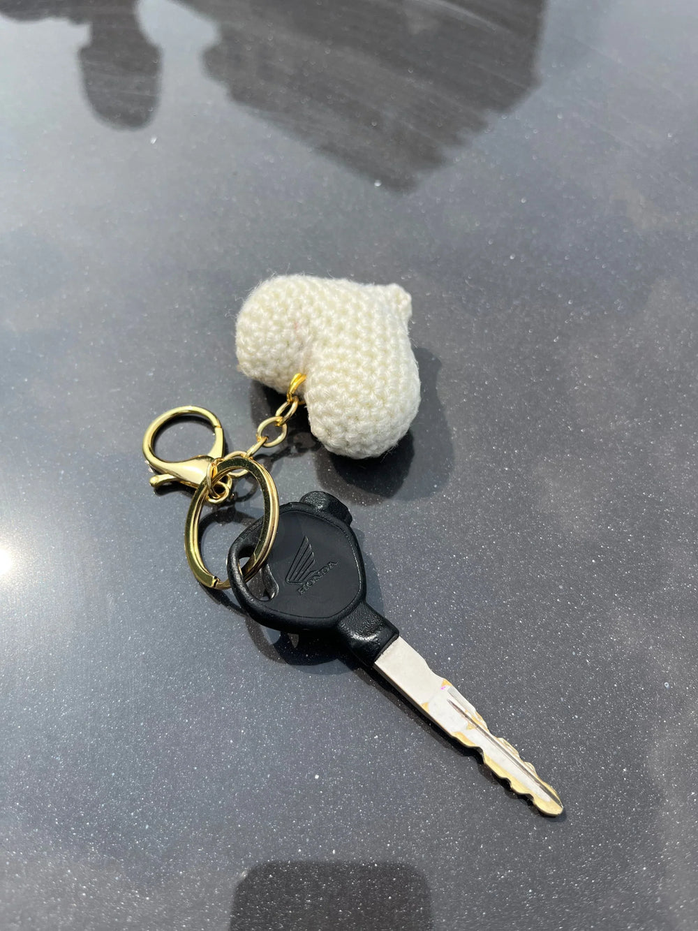 Honda key with gold keychain and white knitted heart charm on dark surface