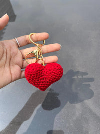 Hand holding a red crochet heart keychain with gold hardware against a grey background