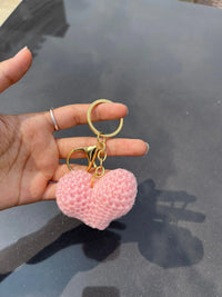 Hand holding a pink crochet heart keychain with gold key ring on a dark background