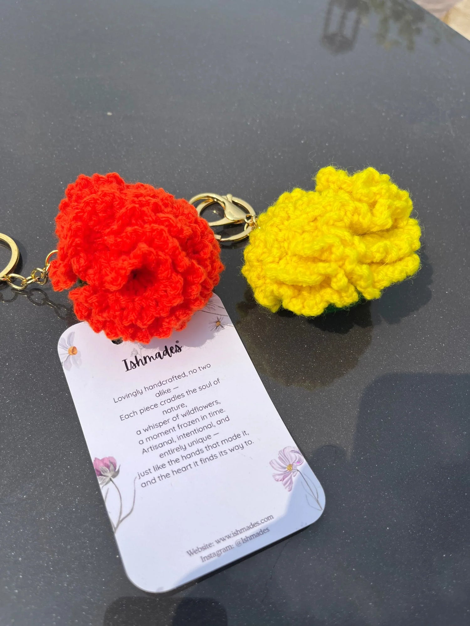 Handcrafted crochet flower keychains in red and yellow with product info card on a dark table.