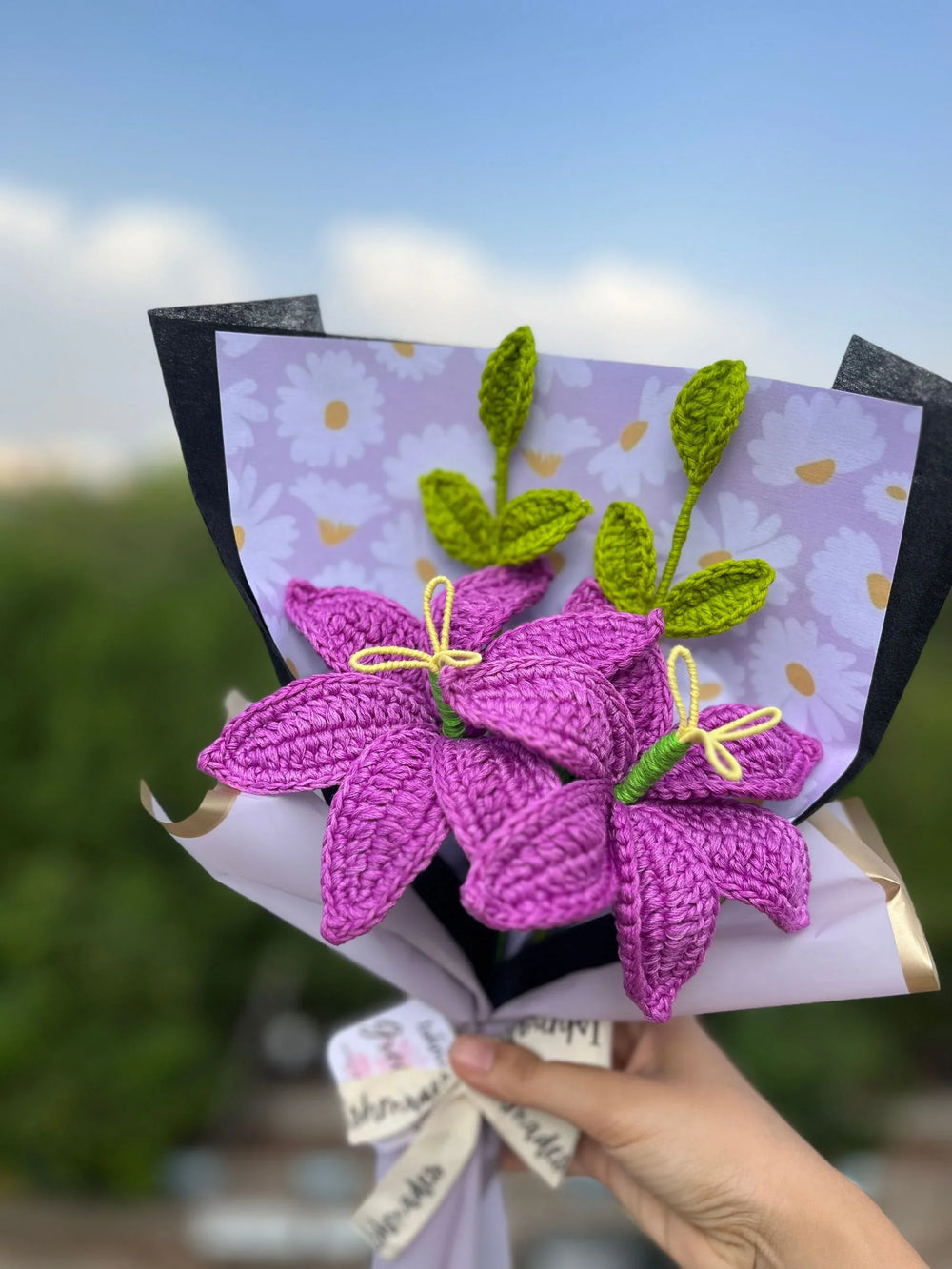 Hand holding bouquet of purple crochet flowers with green leaves, wrapped in floral paper