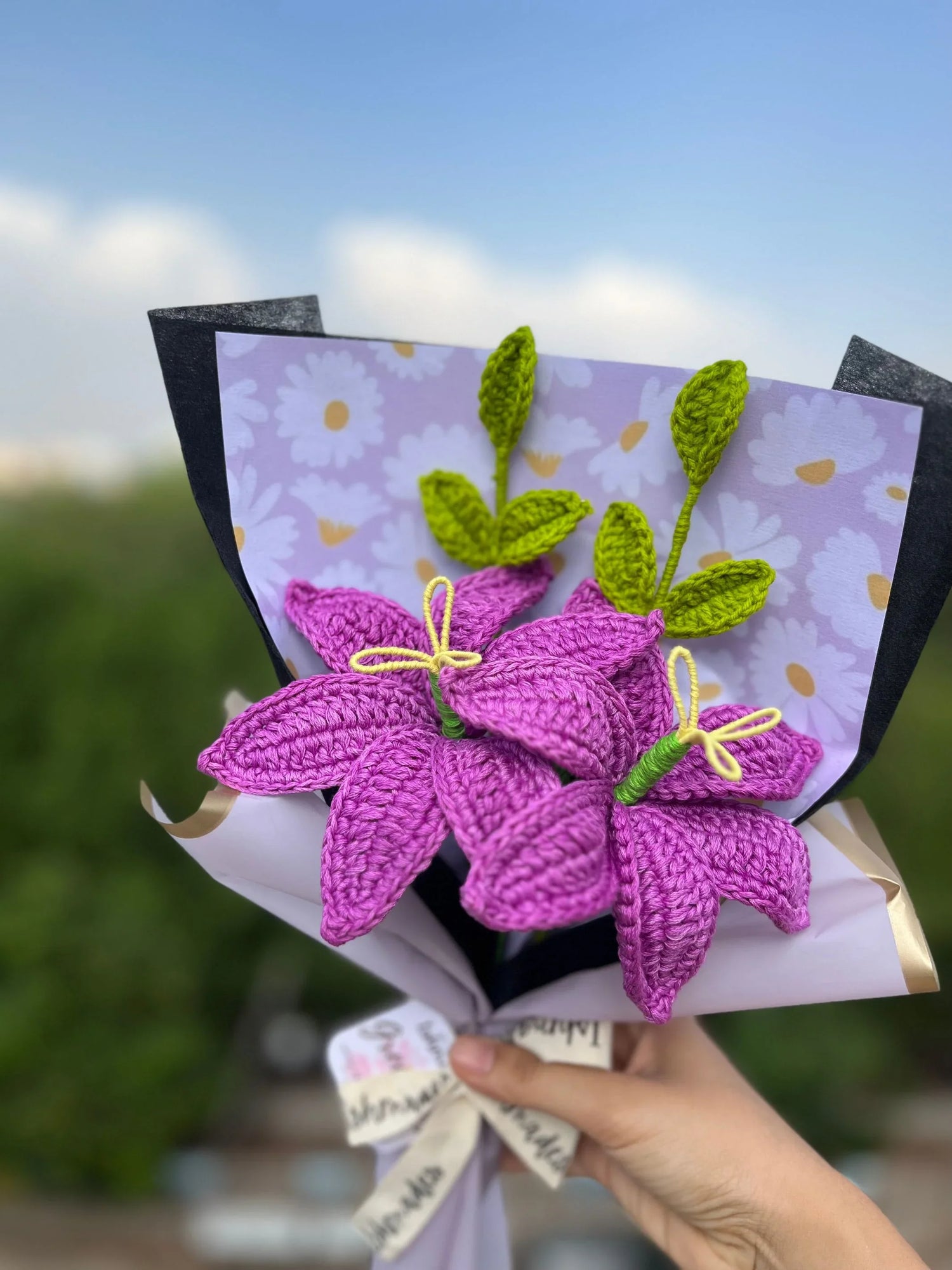 Hand holding bouquet of purple crochet flowers with green leaves, wrapped in floral paper