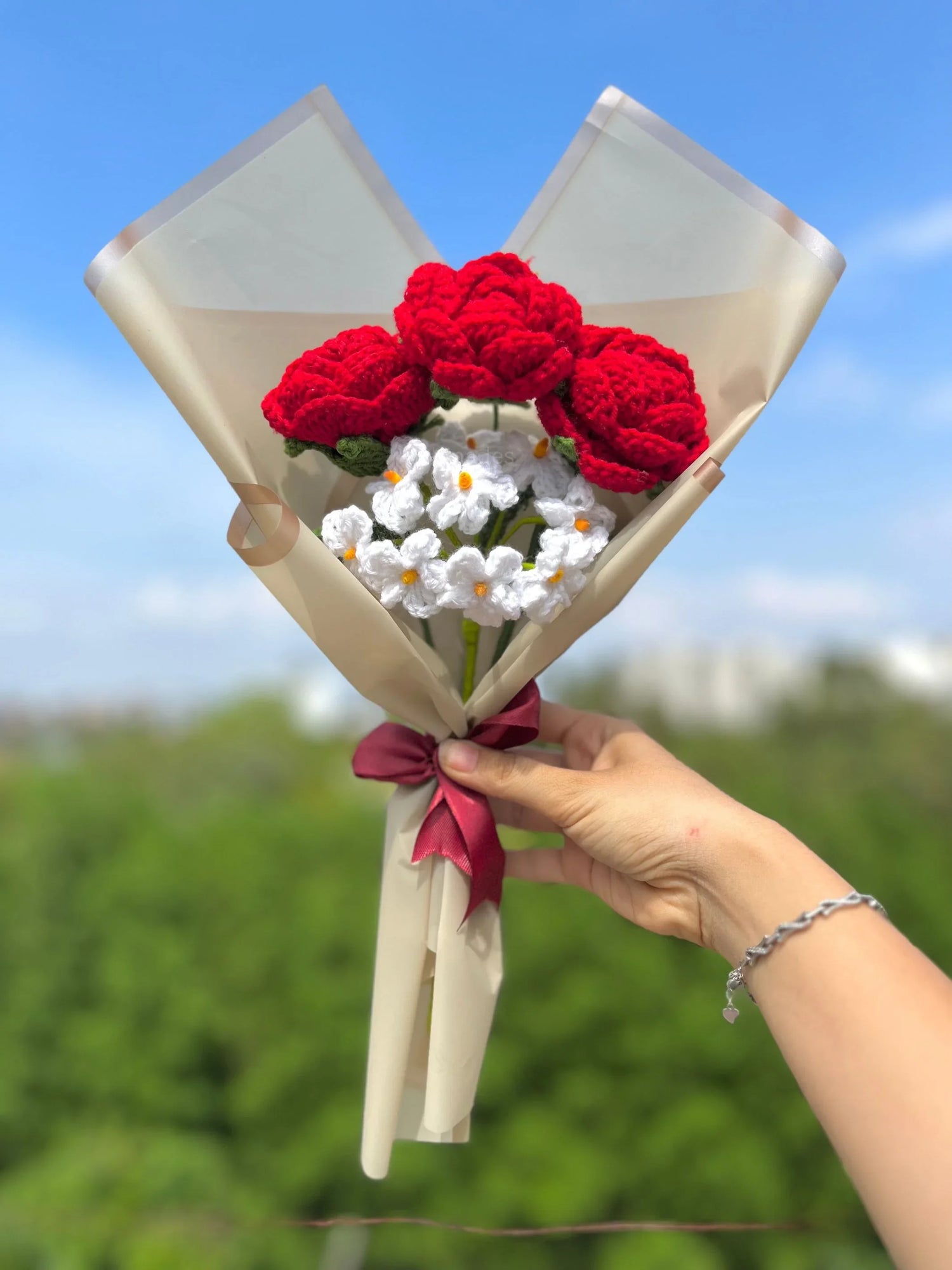 Hand holding crochet flower bouquet with red roses and white daisies against blue sky