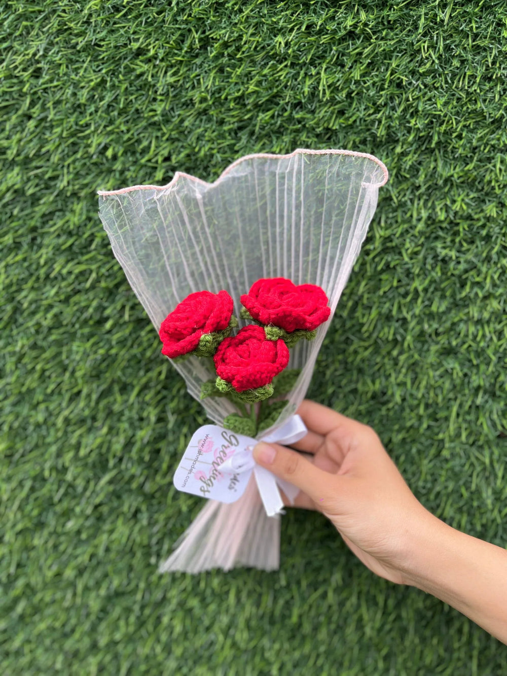 Hand holding bouquet of three red crochet roses wrapped in translucent paper against green grass background