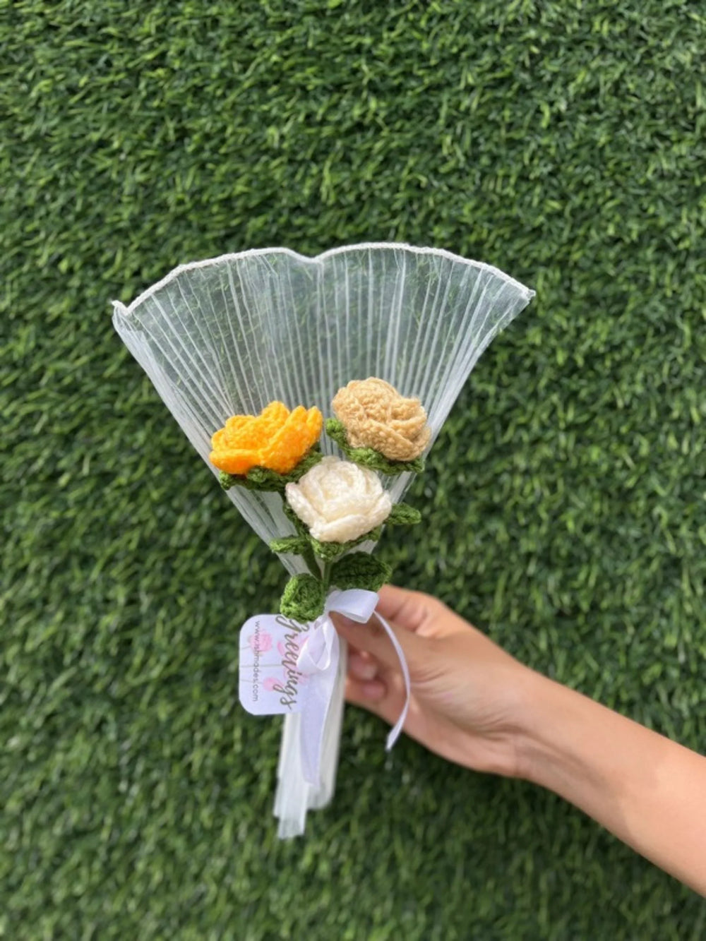 Hand holding a small crochet flower bouquet with white and yellow flowers against a green grass background