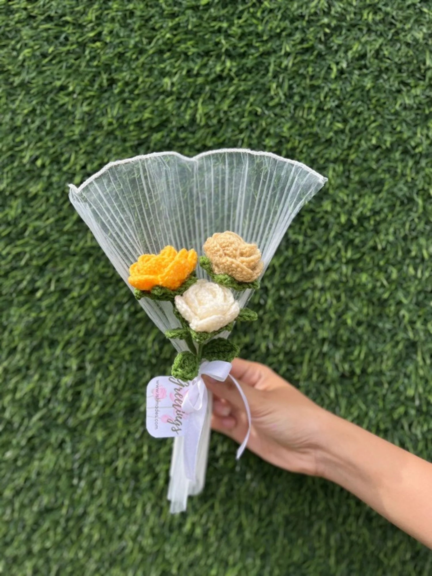 Hand holding a small crochet flower bouquet with white and yellow flowers against a green grass background