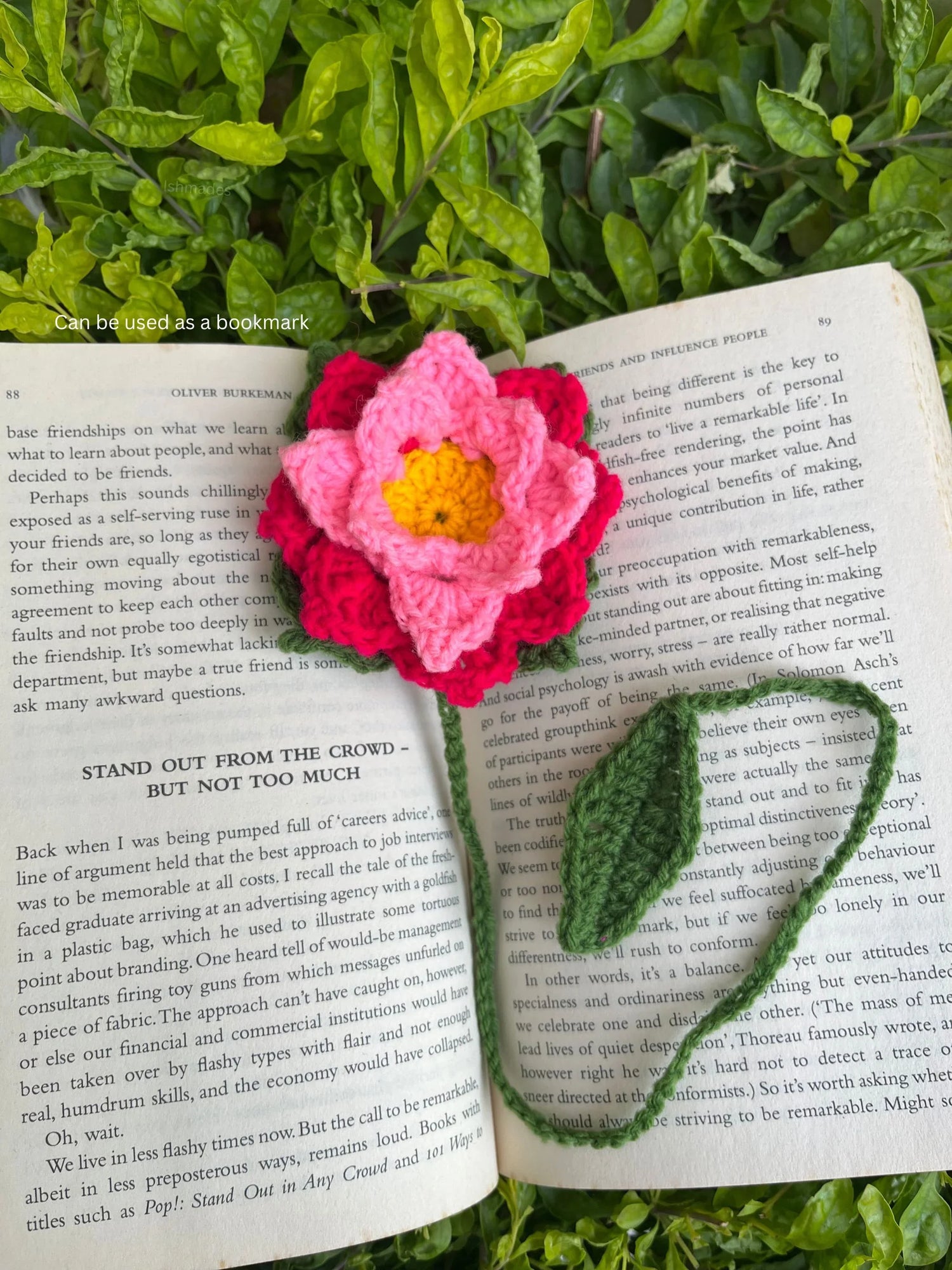 Crochet flower bookmark with pink petals and yellow center on an open book, green leaf stem, outdoors on leafy grass.