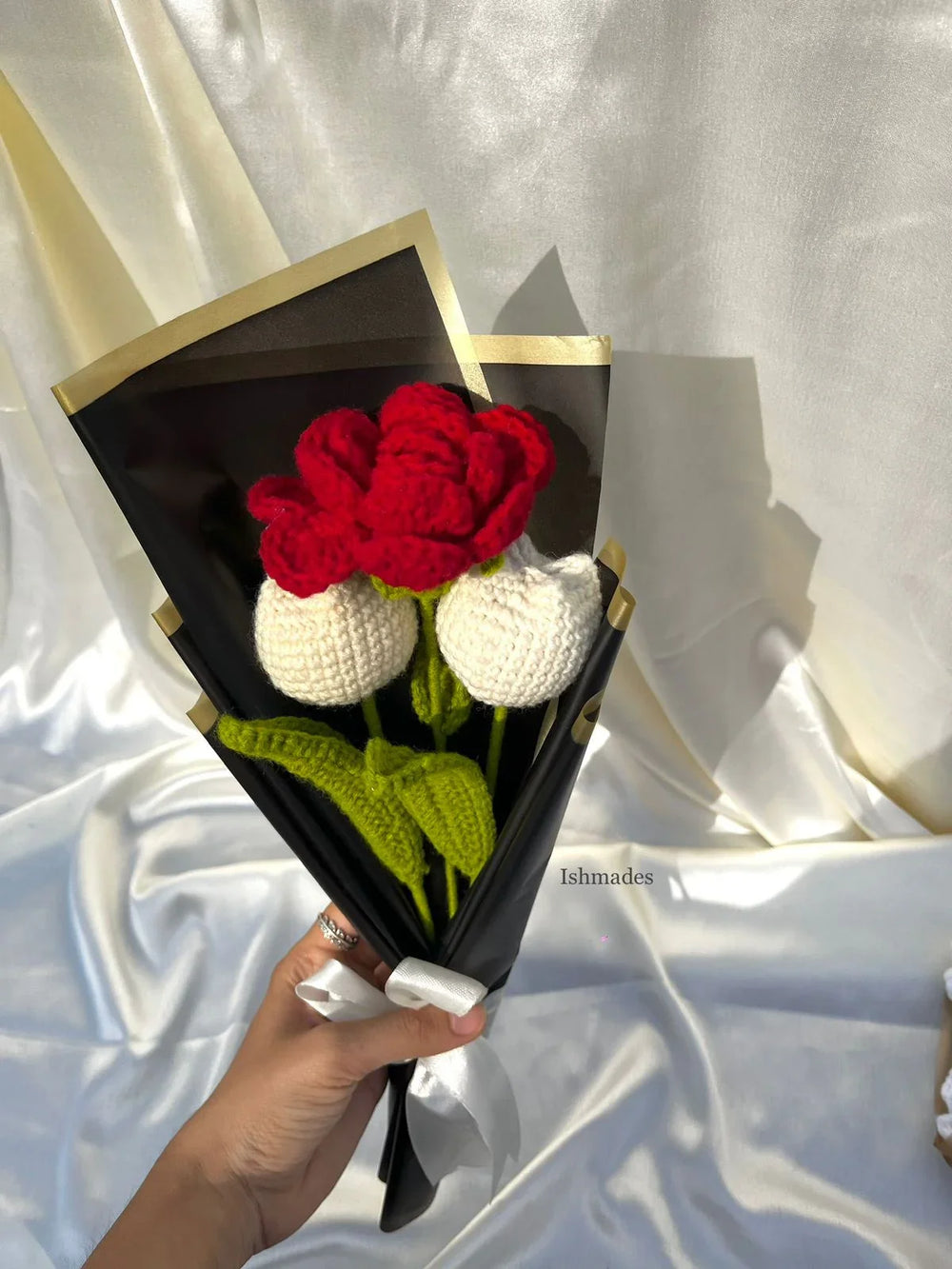 Hand holding a bouquet of crocheted red and white flowers wrapped in black paper with gold edges