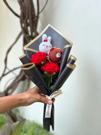 Hand holding a bouquet of red crochet flowers with cute crochet bunny and bear toys, wrapped in black paper.