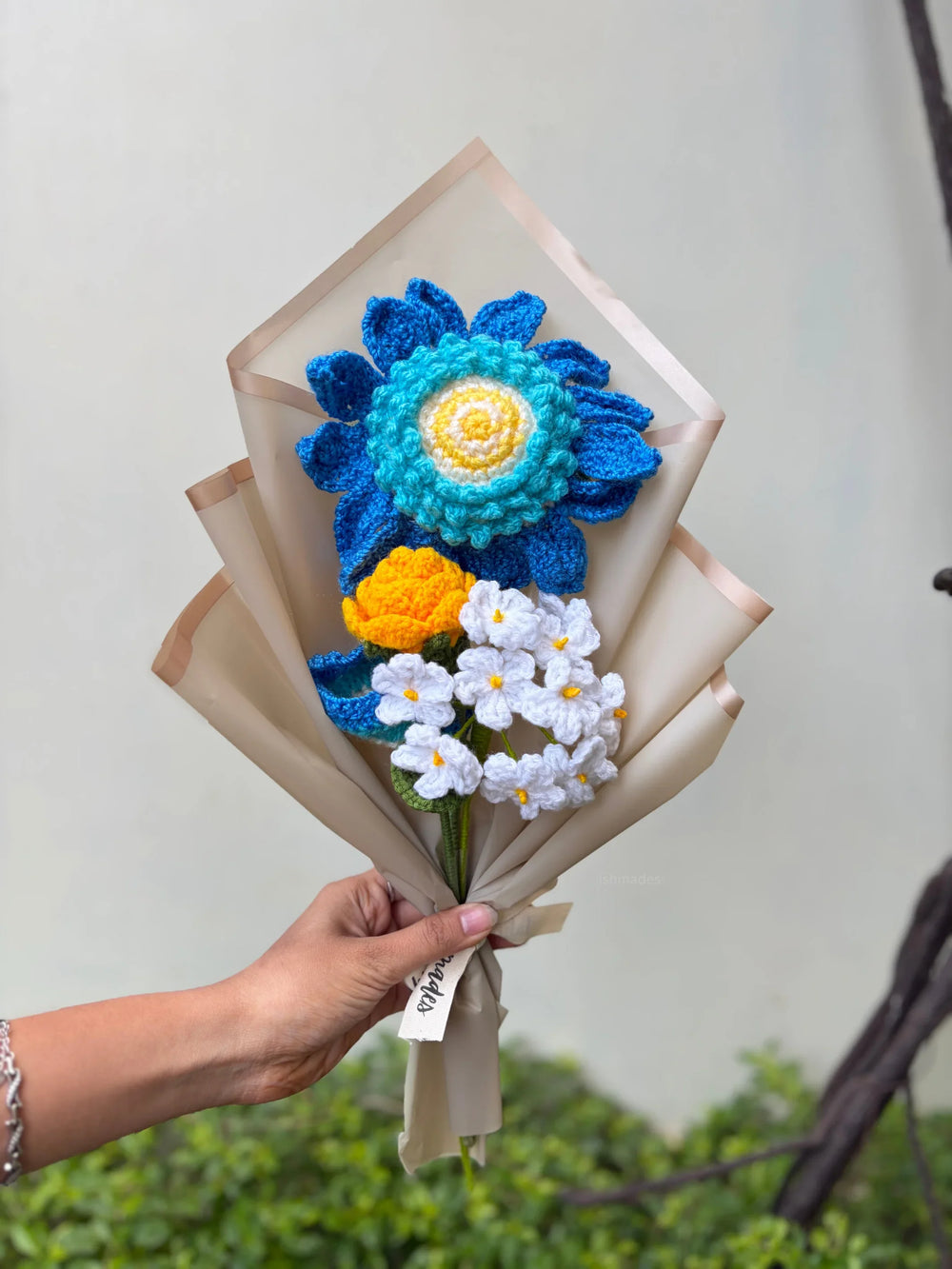 Hand holding a bouquet of colorful crocheted flowers in blue, yellow, and white wrapping paper