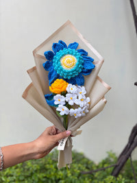 Hand holding a bouquet of colorful crocheted flowers in blue, yellow, and white wrapping paper