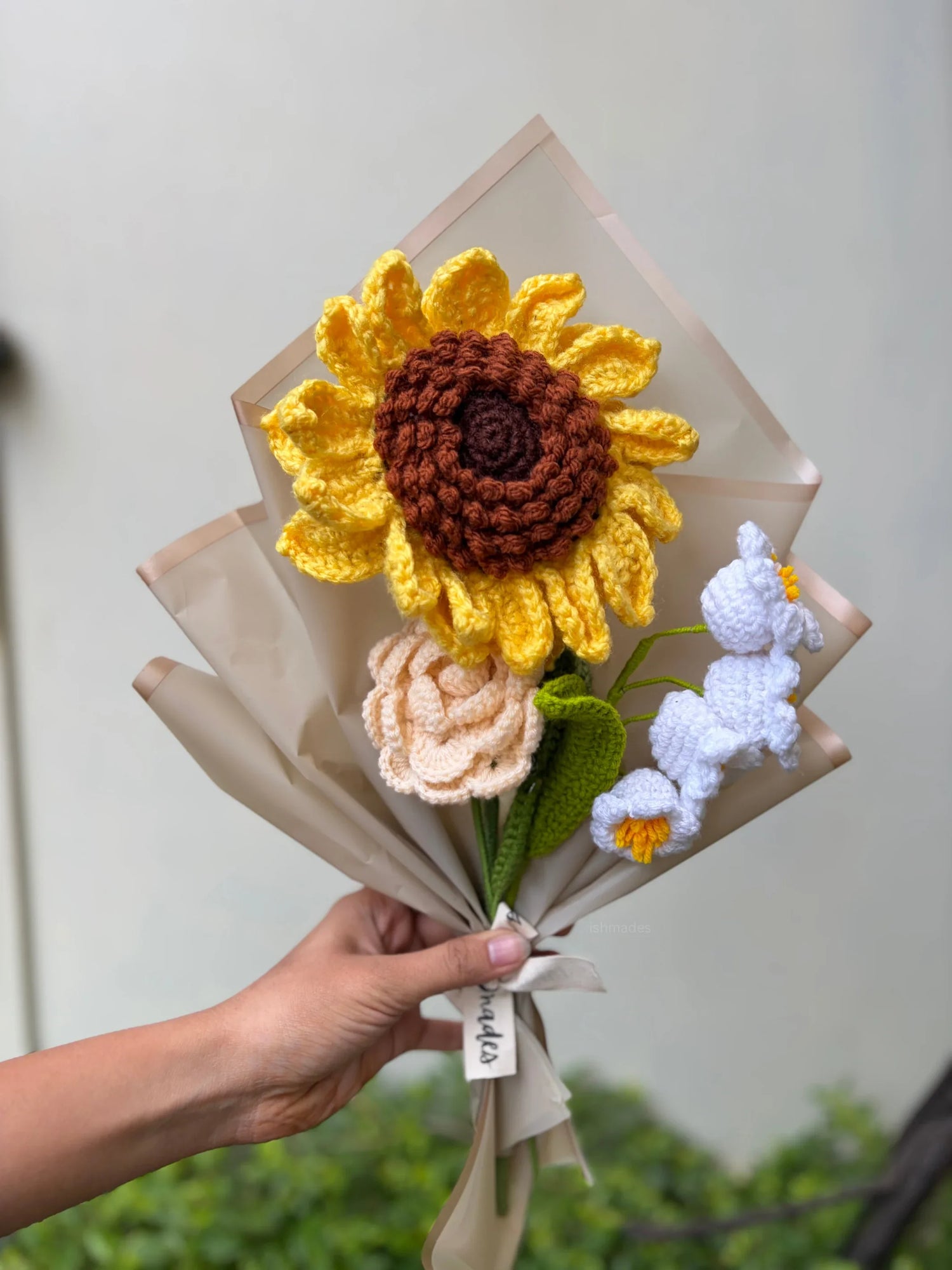 Hand holding a bouquet of knitted flowers including a yellow sunflower and pastel blossoms