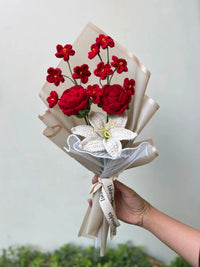 Hand holding a bouquet of crochet flowers with red roses and white lily, wrapped in paper.