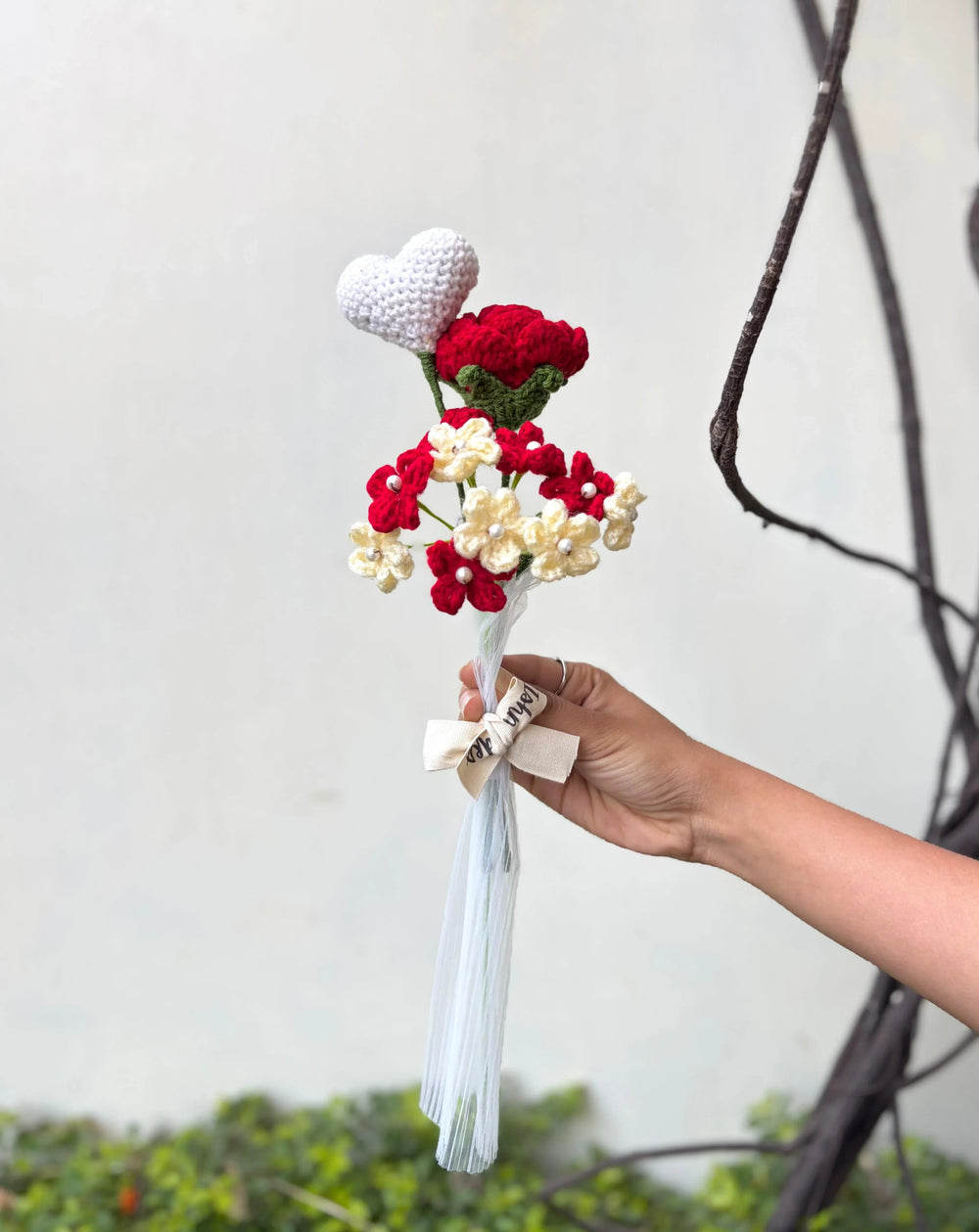 Hand holding crochet bouquet with red flowers, cream flowers, and white heart, tied with a ribbon.
