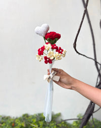 Hand holding crochet bouquet with red flowers, cream flowers, and white heart, tied with a ribbon.