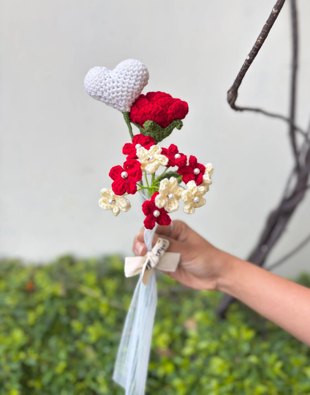 Hand holding crochet bouquet with red and cream flowers, white heart, and ribbon bow