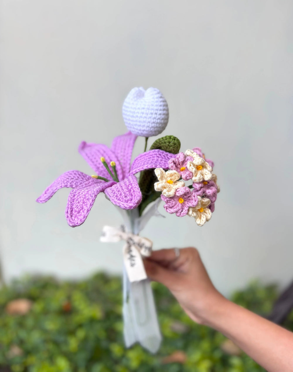 Hand holding crochet flower bouquet with purple lily, white tulip, and pastel blooms