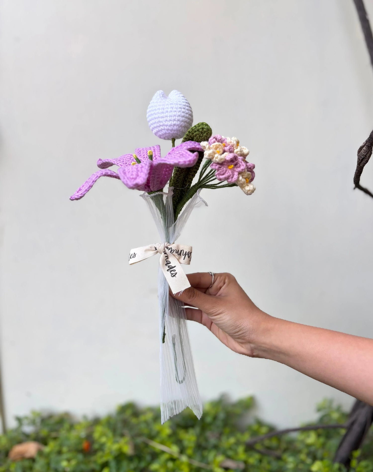Hand holding a bouquet of crocheted purple and pink flowers with a white ribbon outdoors