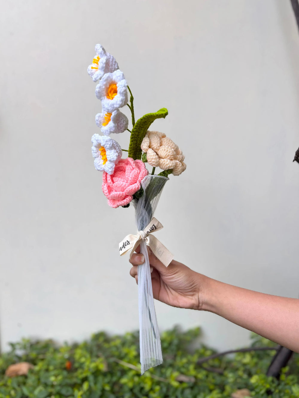 Hand holding a crochet flower bouquet with pink, white, and beige flowers, tied with ribbon.