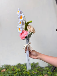 Hand holding a crochet flower bouquet with pink, white, and beige flowers, tied with ribbon.